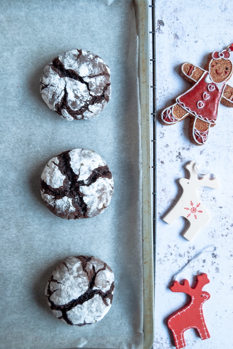 3 chocolate cookies dusted with icing sugar on a silver baking sheet and a gingerbread man and white and red reindeer decorations.