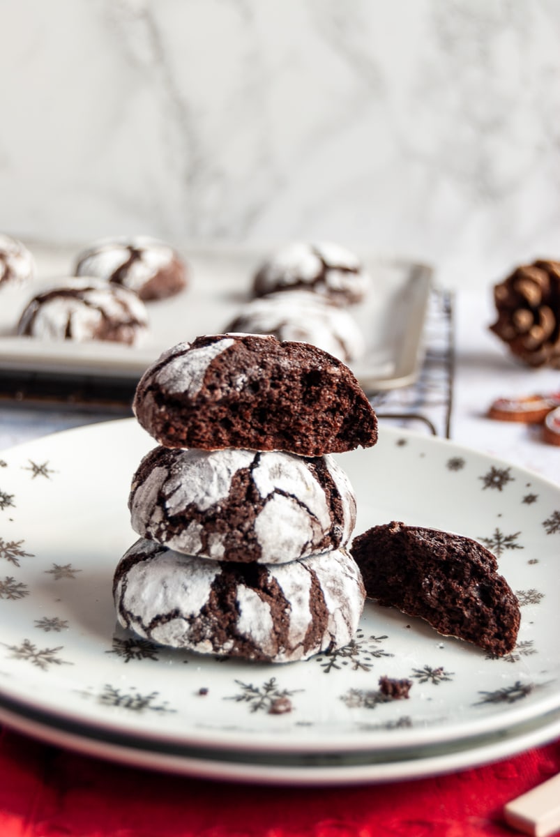 Chocolate cookies dusted with icing sugar on a white and silver star plate with a red napkin. Christmas decorations and more cookies on a baking sheet in the background