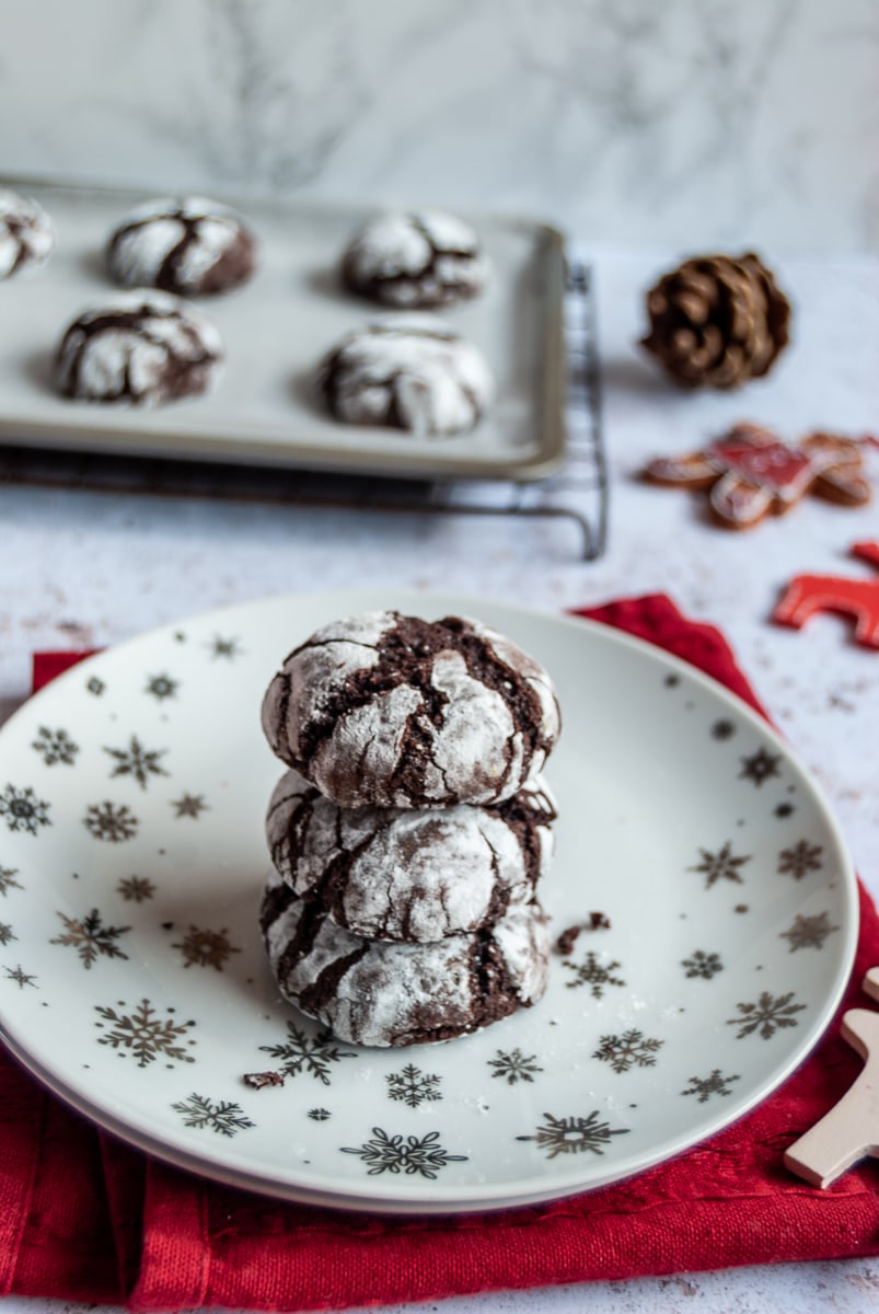 chocolate fudge cookies on a white and silver star plate, a red napkin, red and white reindeer decorations and more cookies on a baking sheet.