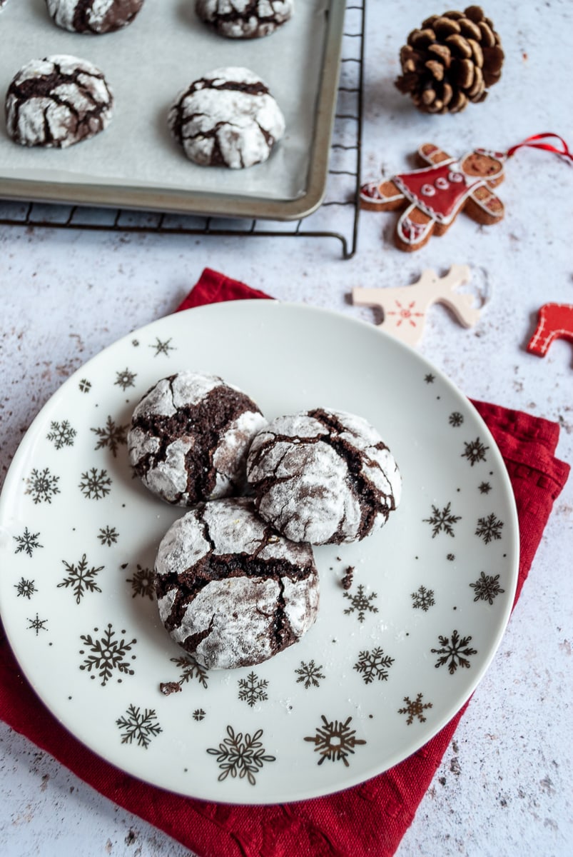 chocolate crinkle cookies on a white and silver star plate, a red napkin, Reindeer and gingerbread men decorations and more cookies on a baking sheet.