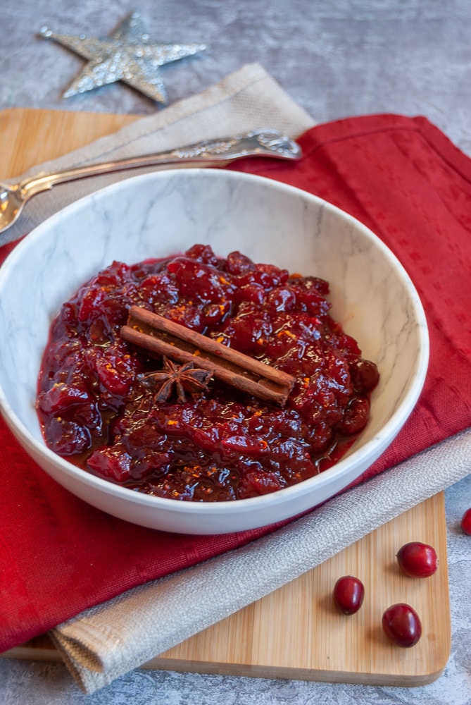 A white bowl of cranberry sauce topped with a cinnamon stick and star anise sitting on a red napkin with a silver serving spoon. 