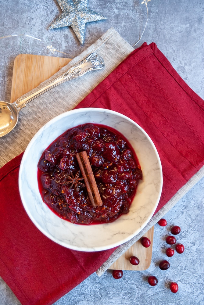 A flat lay photo of a white bowl of cranberry sauce sitting on red and beige napkins, a silver serving spoon and fresh cranberries 