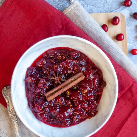 A white bowl of cranberry sauce topped with a cinnamon stick on a red napkin.