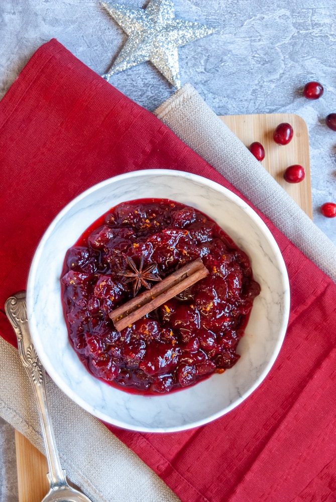 A bowl of cranberry sauce topped with a cinnamon stick and a star anise on red and beige napkins, a silver spoon and fresh cranberries with a silver star decoration 