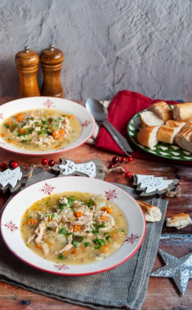 two red and white bowls of turkey, vegetable and rice soup with grey and napkins, wooden salt and pepper pot and a green plate of bread and soup spoons.