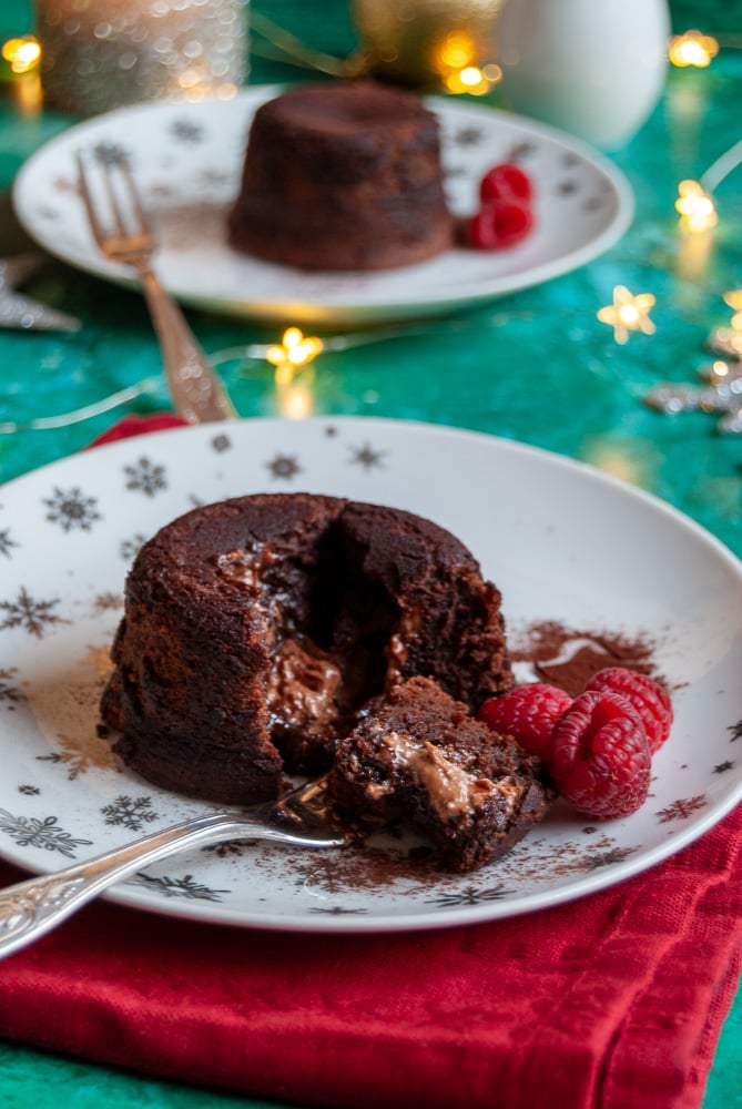 a mini chocolate pudding cut in half on a white and silver plate with raspberries, a red napkin and another pudding on a white plate in the background with fairy lights.