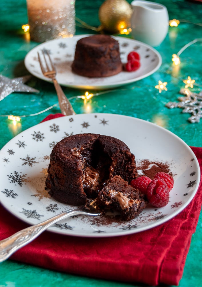 two mini chocolate puddings with raspberries and a small fork on a white and silver plates, a red napkin and gold fairy lights