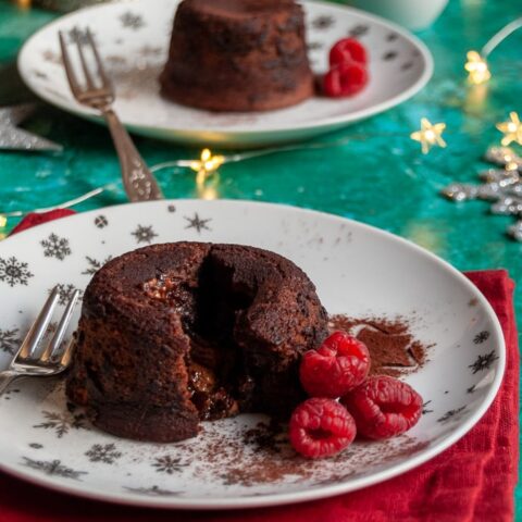 two mini chocolate puddings with raspberries on on white a silver plate and red napkin, gold fairy lights and a white jug and candle