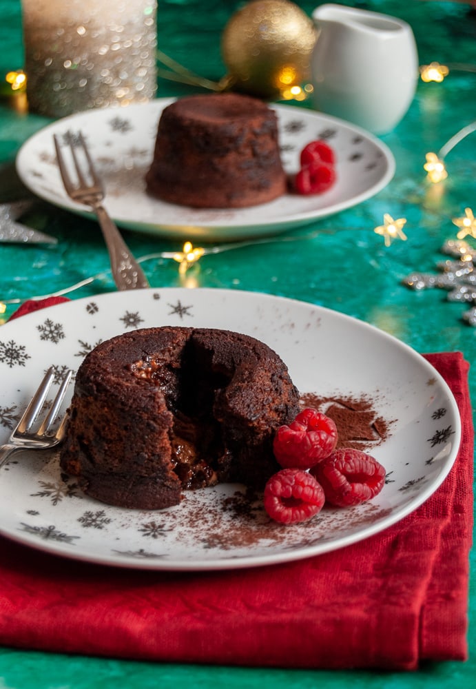 two mini chocolate puddings with raspberries on on white a silver plate and red napkin, gold fairy lights and a white jug and candle