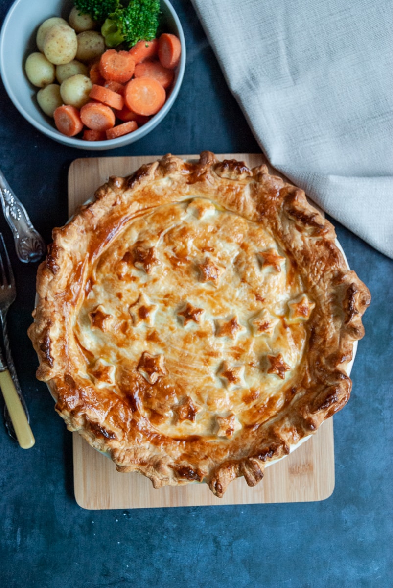 A creamy turkey and ham pie on a wooden chopping board and a bowl of carrots, potatoes and broccoli. 