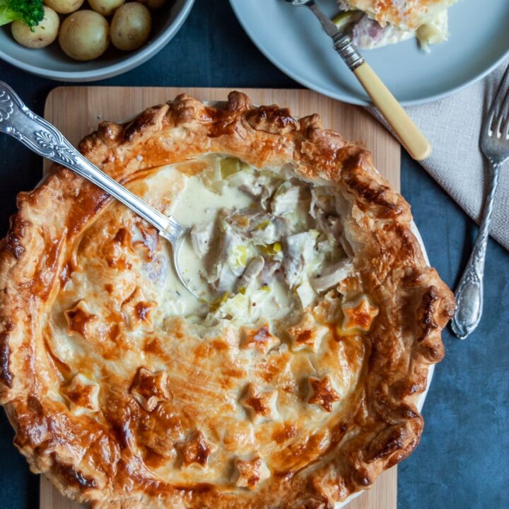 a flat lay photo of a turkey and ham pie with a spoon digging inside, revealing the creamy filling, a bowl of potatoes, carrots and broccoli and a small plate with a piece of pie and a fork.