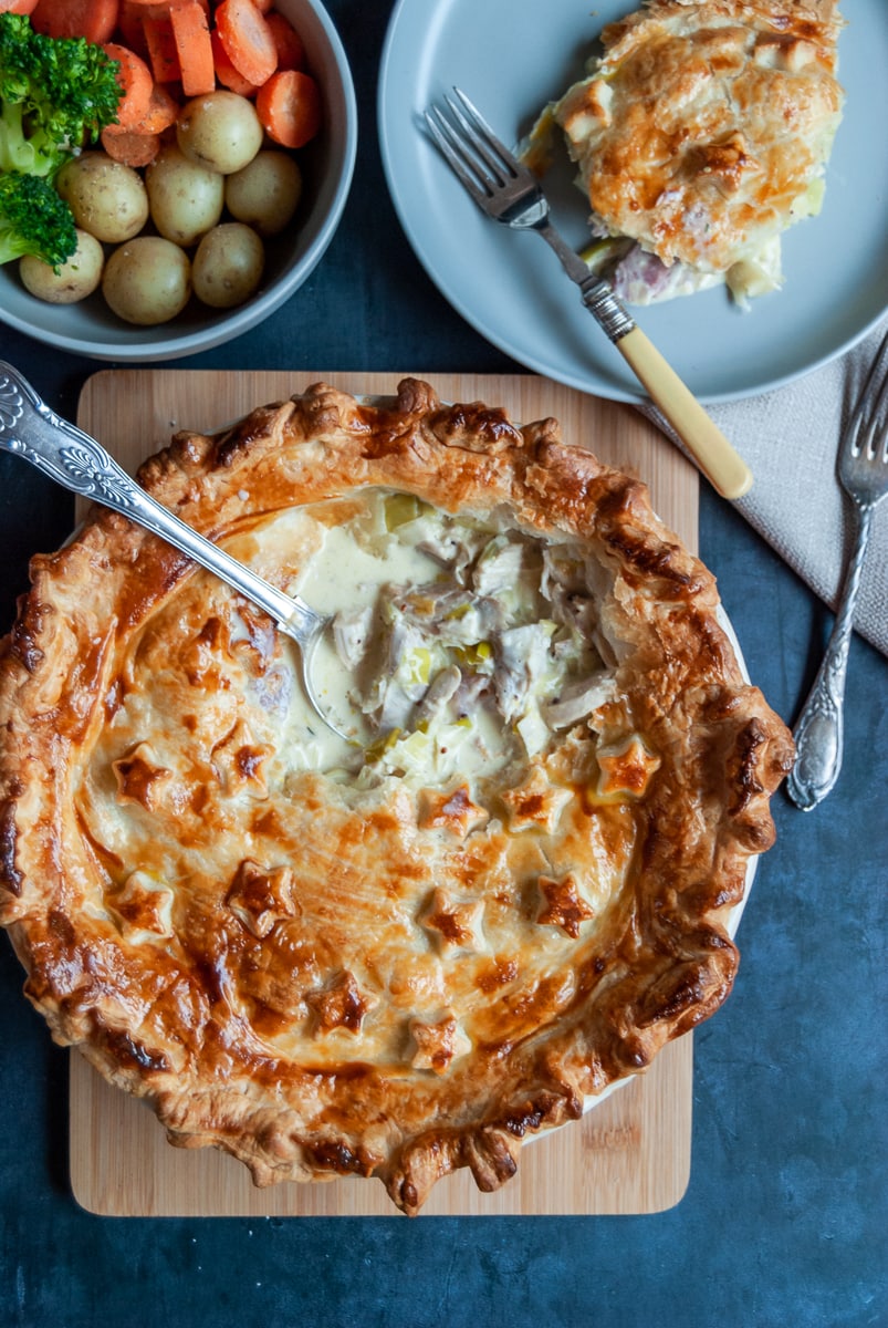 a flat lay photo of a turkey and ham pie with a spoon digging inside, revealing the creamy filling, a bowl of potatoes, carrots and broccoli and a small plate with a piece of pie and a fork.