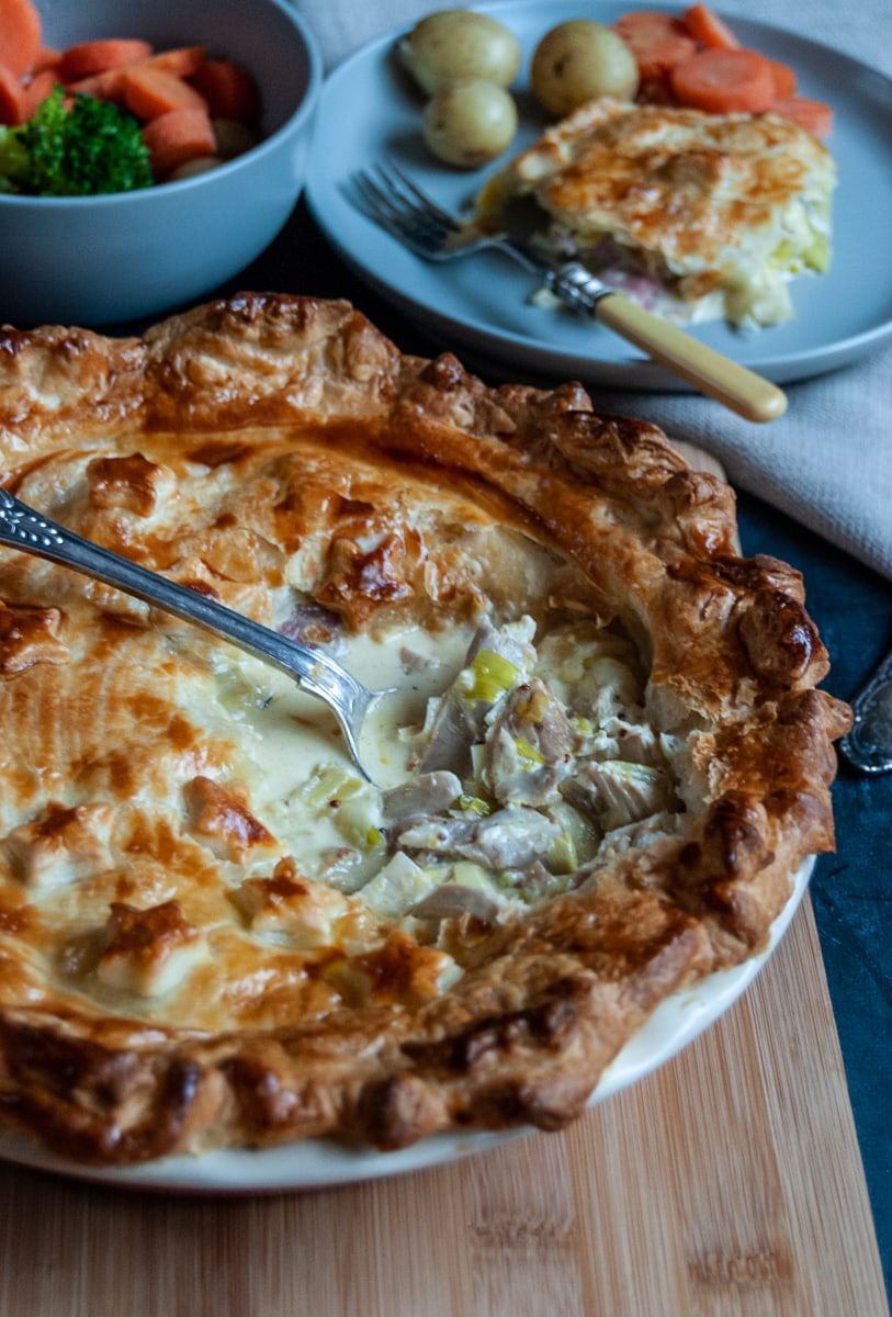 a turkey and ham pie with a spoon digging in to reveal the creamy filling, a bowl of broccoli and carrots and a small plate with a slice of the pie, potatoes and a fork.