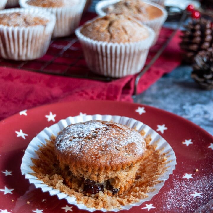 A mincemeat muffin dusted with icing sugar on a red and white star plate. More muffins siting on a black wire rack and red tablecloth in the background.