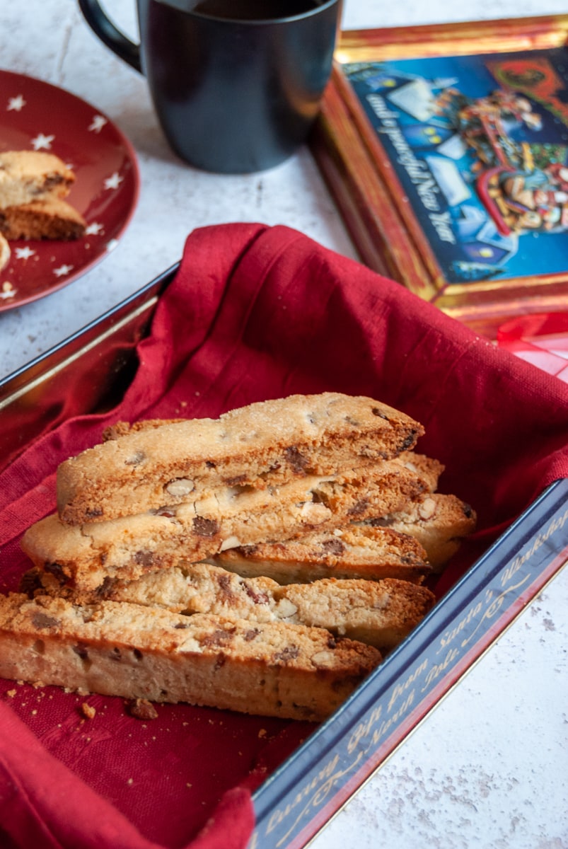 a box of chocolate chip and nut biscotti in a box lined with a red napkin, a black mug of coffee and a red and white star plate can be partially seen.