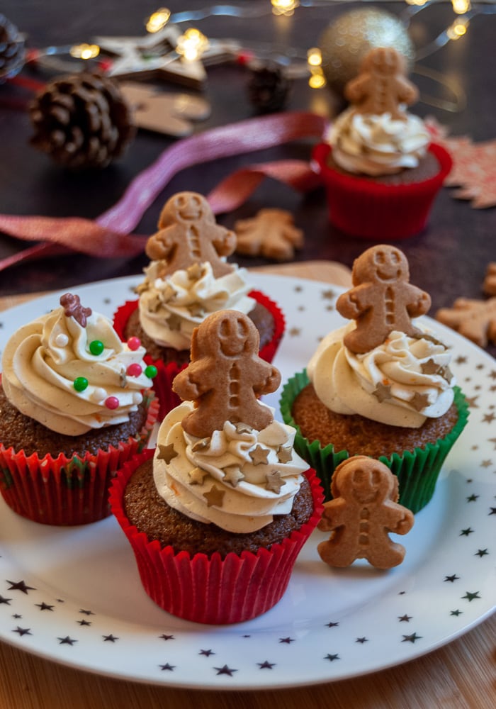 ginger cupcakes on a white and gold star plate topped with orange buttercream, Christmas sprinkles and mini gingerbread men.  Fairy lights and Christmas ribbon in the background.