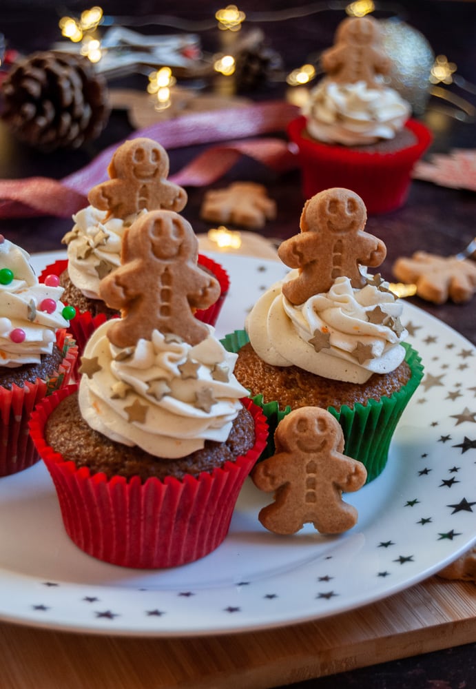 gingerbread cupcakes on a white and gold star plate decorated with orange buttercream swirls, mini gingerbread people and spinkles.  Fairy lights and Christmas ribbon is in the background