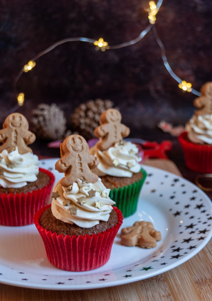three ginger cupcakes on a white and gold star plate topped with orange buttercream swirls and a mini gingerbread person with fairy lights in the background