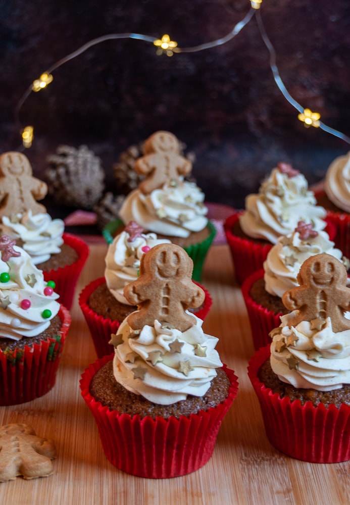 gingerbread cupcakes on a wooden board decorated with orange buttercream swirls and topped with a mini gingerbread person.  Fairy lights can be seen in the background