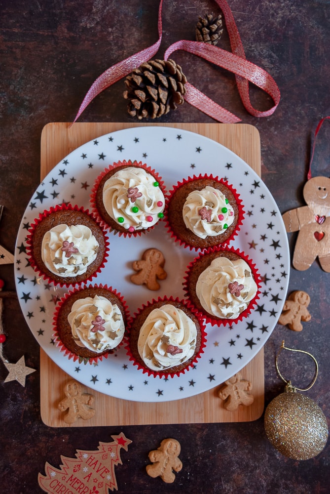 a flat lay photo of a plate of ginger cupcakes topped with orange buttercream and gold sprinkles. Mini gingerbread people and red and gold Christmas decorations have been placed around the plate.