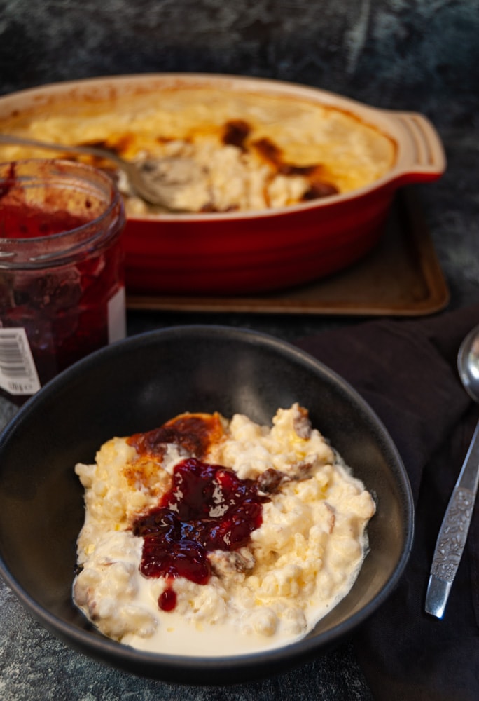 A black of rice pudding with a jar of jam in the background and a dish of rice pudding with a large silver serving spoon.