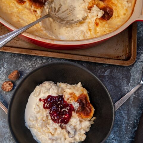 a black stoneware dish of rice puding with sultanas and topped with raspberry jam and a red and white dish of the pudding with a vintage spoon on a metal baking tray.