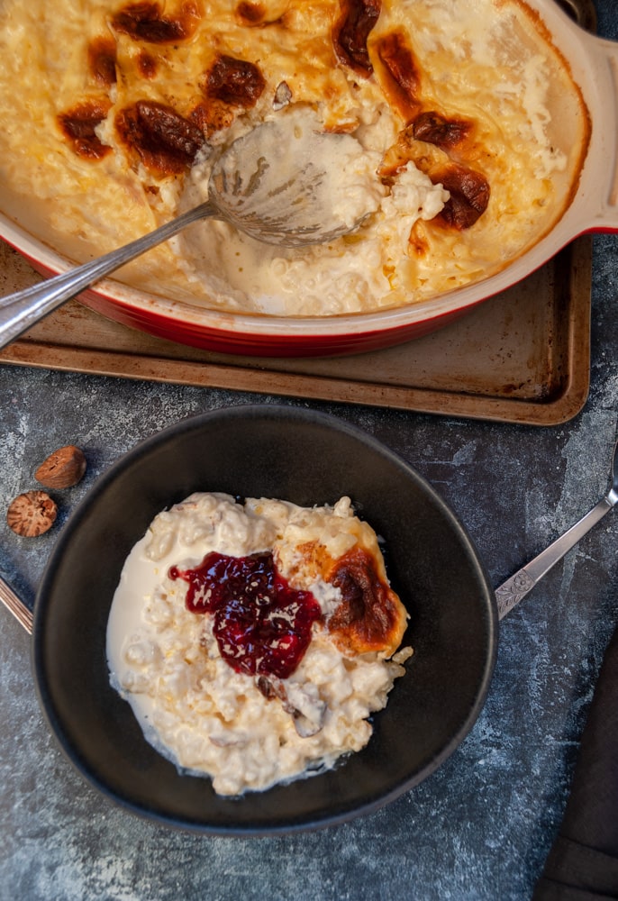 a black stoneware dish of rice puding with sultanas and topped with raspberry jam and a red and white dish of the pudding with a vintage spoon on a metal baking tray.