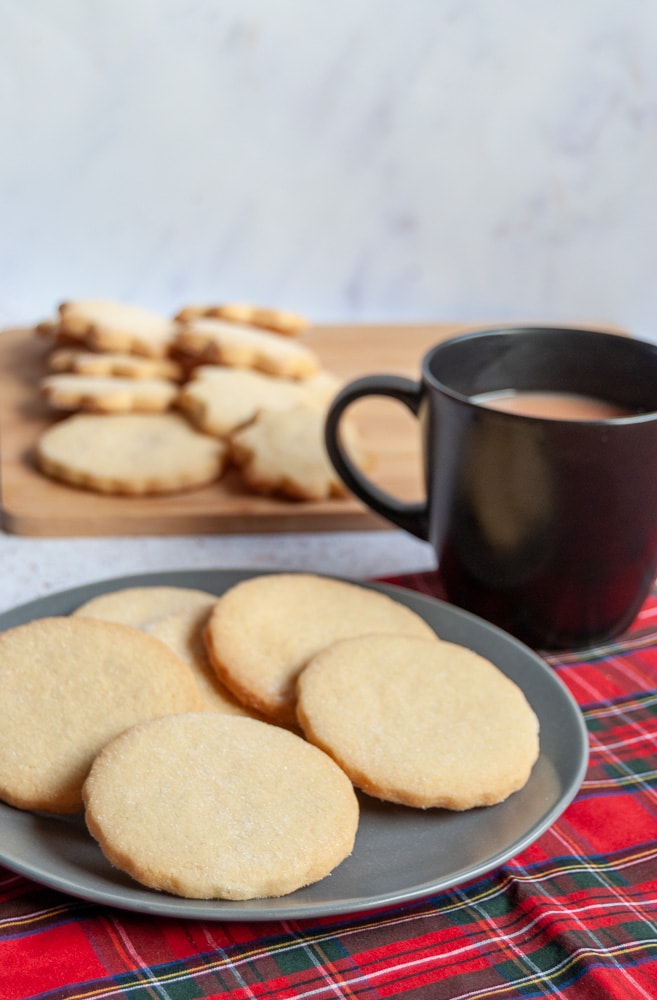 a grey plate with pieces of shortbread on a tartan tablecloth, a black mug of tea and a wooden board with more pieces of shortbread.