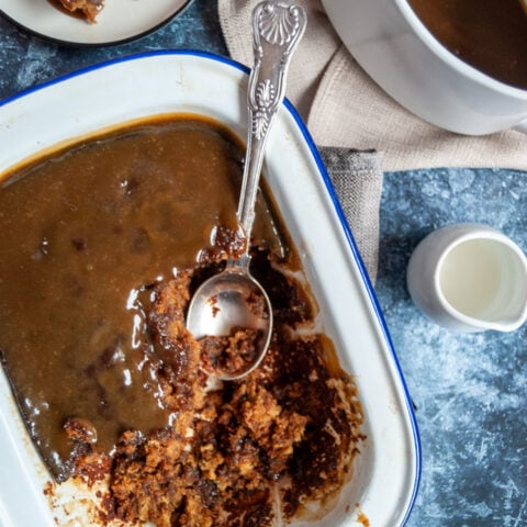 a white and blue enamel dish of toffee pudding with a large silver spoon, a small plate with a piece of the pudding, a large white jug of toffee sauce and a small white jug of cream on a blue and white background.