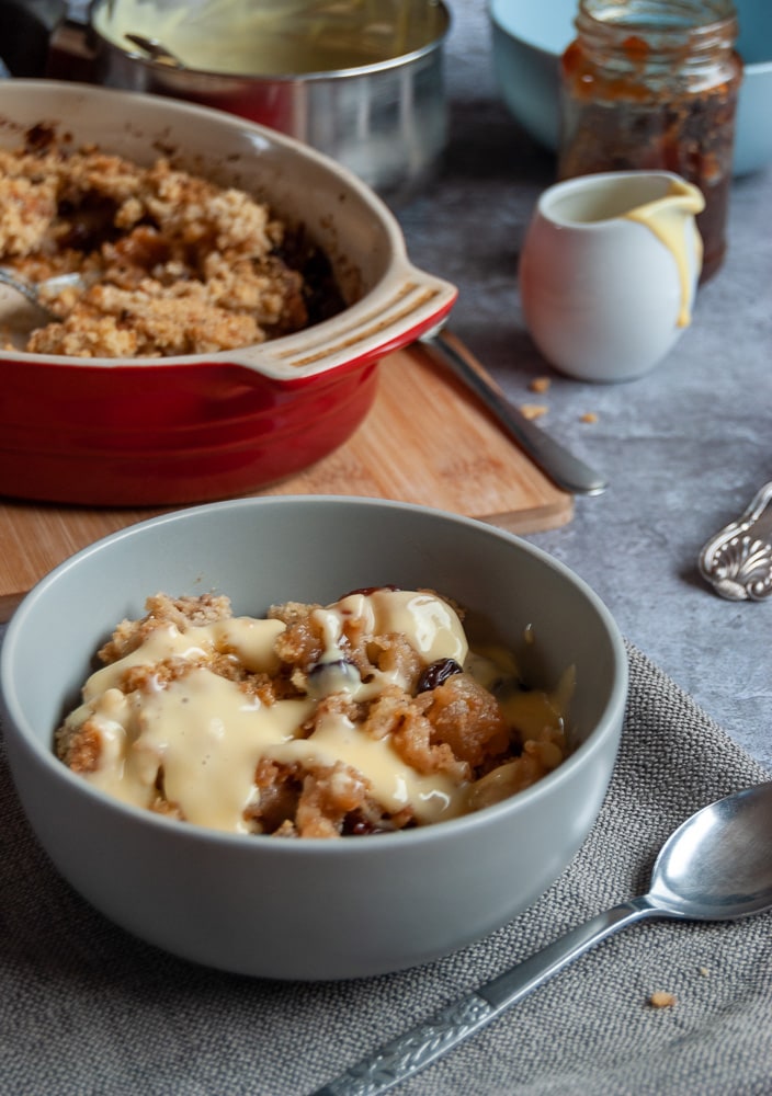 a light blue bowl of apple and mincemeat crumble on a grey napkin and a silver spoon, a red dish of the crumble, a small jug of custard and a jar of mincemeat