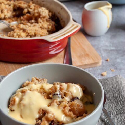 a bowl of apple and mincemeat crumble on a grey napkin, an oval red dish of crumble and a small white jug of custard in the background.