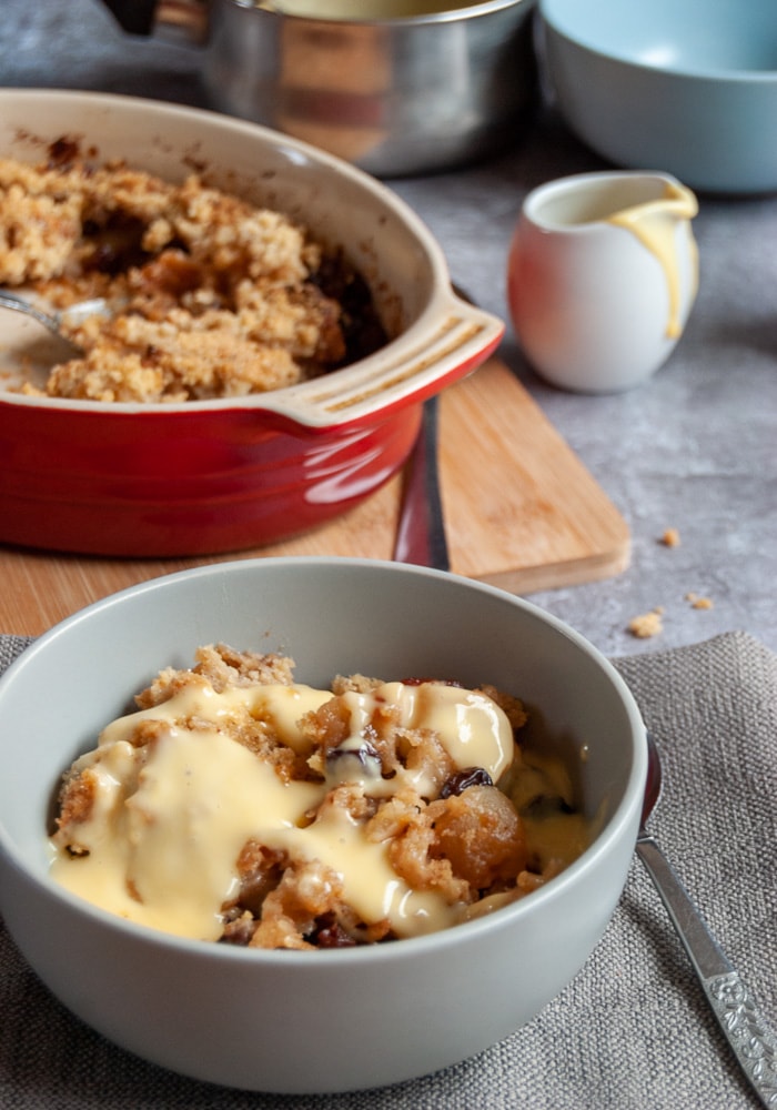 a bowl of apple crumble with mincemeat on a grey napkin, an oval red dish of crumble and a small white jug of custard in the background.