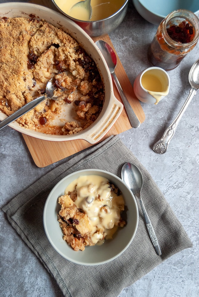 a flatlay photo of a bowl of apple crumble with custard, an oval dish of crumble with a sliver spoon and a small jug and pan of custard