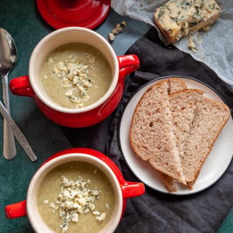 two red bowls of broccoli and stilton soup, a plate of brown bread on a black napkin, two silver spoons and a piece of greaseproof parchment with a piece of stilton cheese.