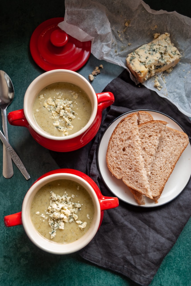 two red bowls of broccoli and stilton soup, a plate of brown bread on a black napkin, two silver spoons and a piece of greaseproof parchment with a piece of stilton cheese.