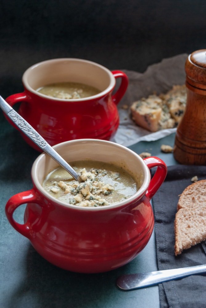 two red bowls of broccoli soup topped with crumbled stilton cheese, a wooden pepper mill and a piece of brown bread on a black napkin.