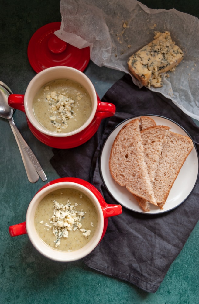 two red bowls of broccoli and stilton soup, a plate of brown bread on a black napkin, two silver spoons and a piece of greaseproof parchment with a piece of stilton cheese on a dark green background.