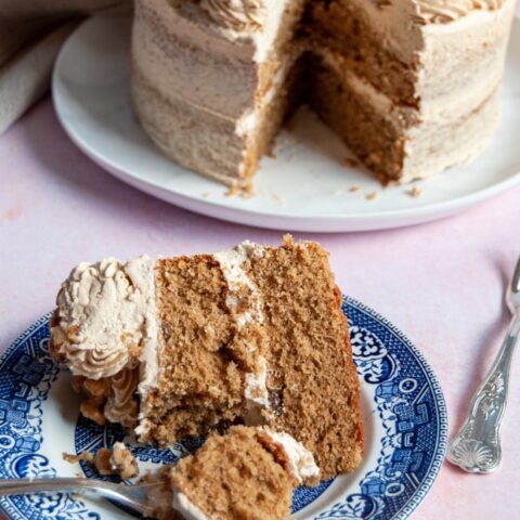 a slice of coffee and walnut cake with a silver cake fork on a blue willow patter cake and a large coffee cake with a slice removed on a white plate on a light pink backdrop.