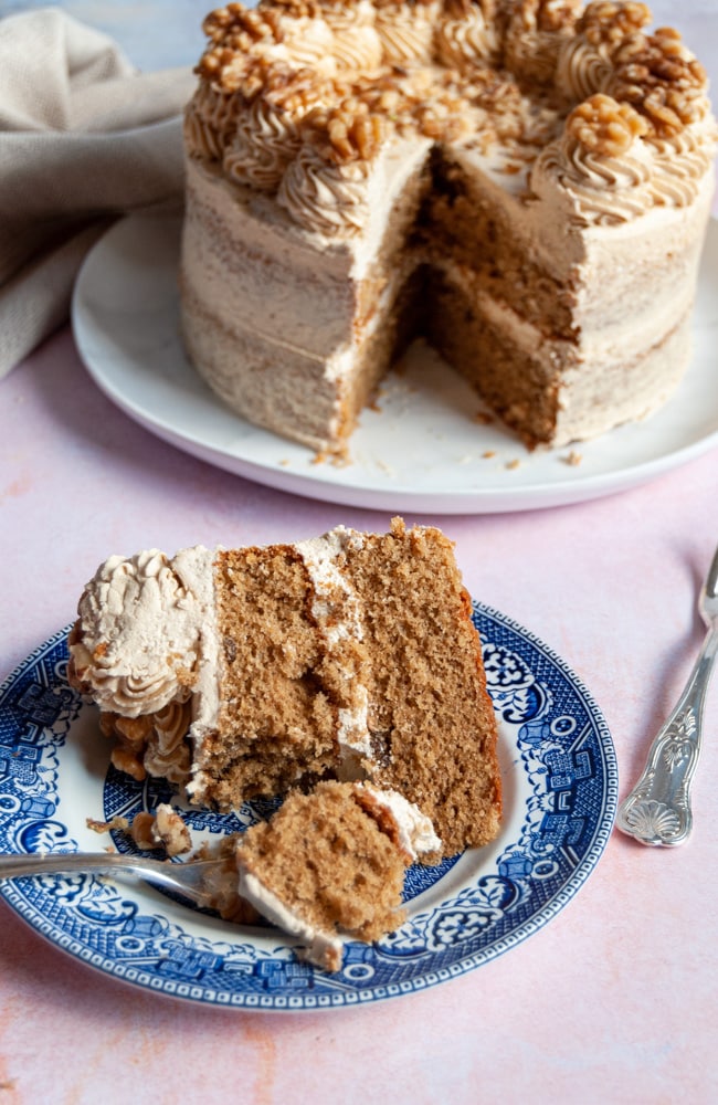 a slice of a large coffee cake decorated with coffee buttercream and walnuts with a silver cake fork on a blue willow patter cake and a large coffee cake with a slice removed on a white plate on a light pink backdrop.