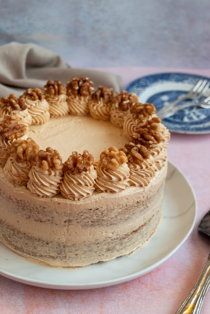 A coffee layer cake decorated with coffee buttecream frosting and topped with whole walnuts on a white plate on a pink backdrop.  A blue Willow Pattern plate and a vintage cake slice can be partially seen beside the cake.