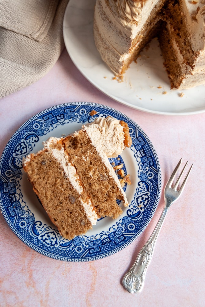 a slice of coffee cake with coffee buttercream on a blue willow pattern plate, a silver cake fork and a large coffee cake with a slice removed on a white plate and light pink backdrop.