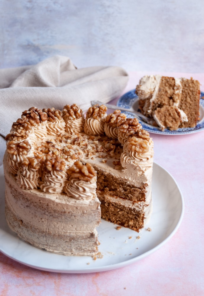 a coffee cake with a slice removed to reveal the fluffy interior on a white plate.  The cake is topped with coffee buttercream swirls and whole walnuts.  A slice of the cake on a blue and white plate with a cake fork and a beige linen napkin is in the background.