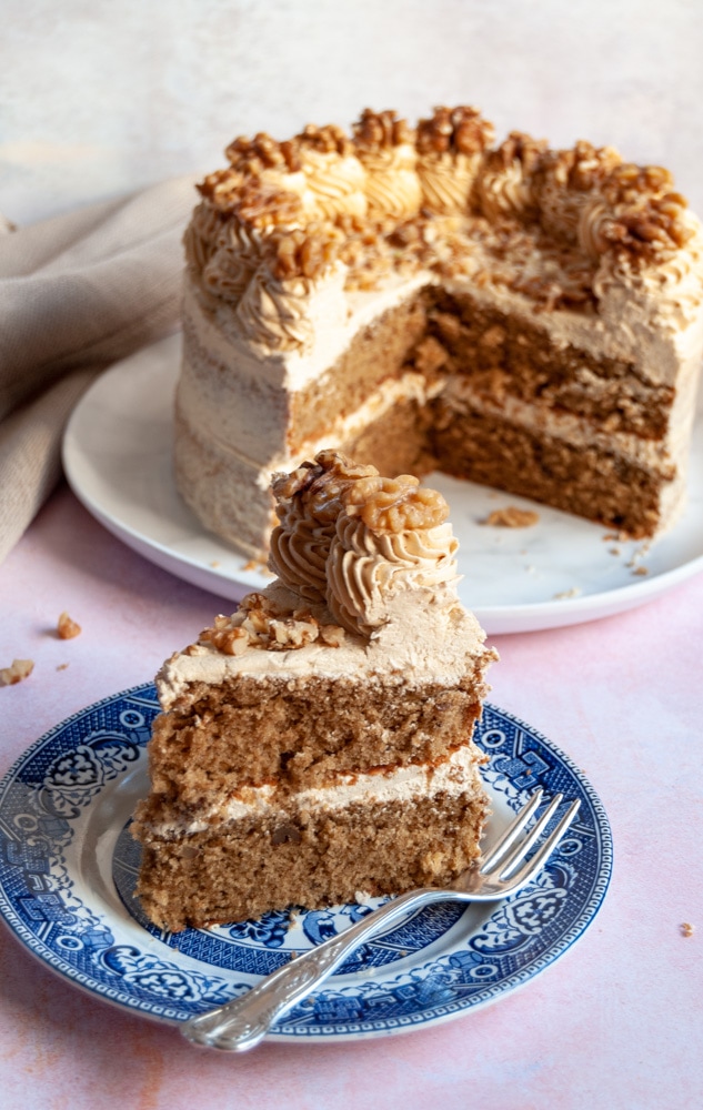 a slice of coffee cake topped with coffee buttercream and a walnut on a blue and white plate with a silver cake fork.  A large coffee cake with slices removed from it is sitting in the background on a white plate.