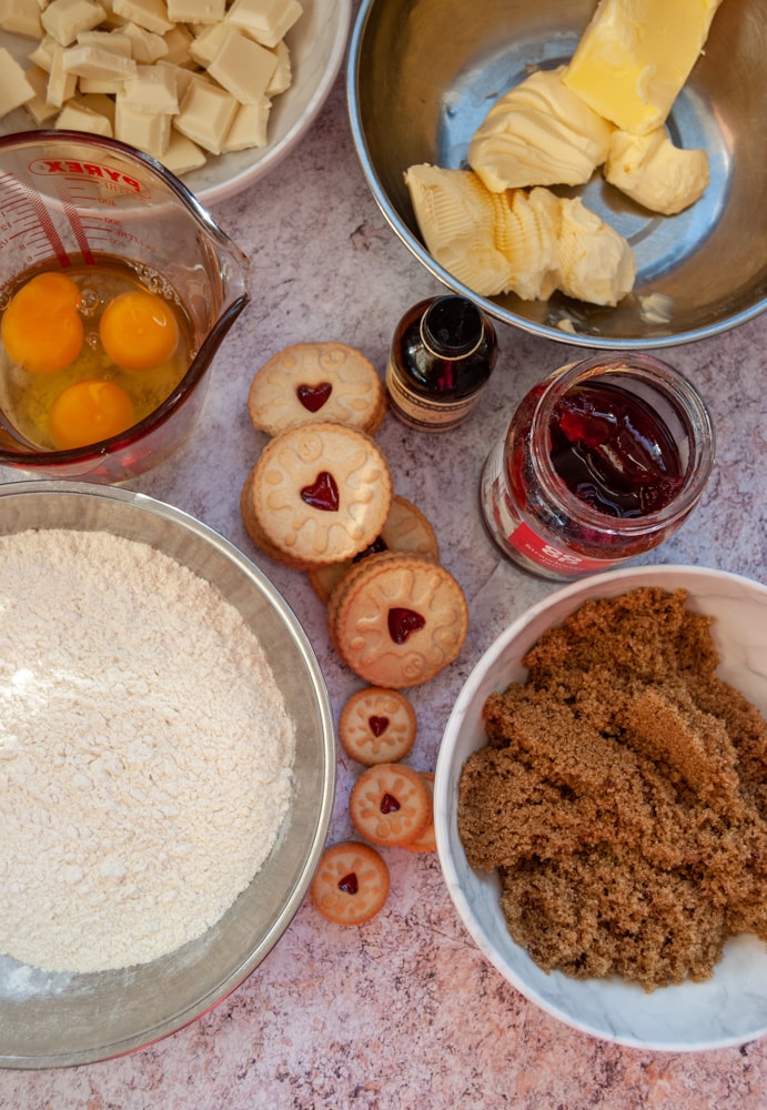 bowls of brown sugar, flour, butter, white chocolate, eggs, vanilla extract and a jar of jam on a grey worktop.