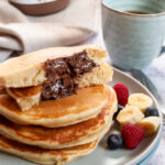 a stack of pancakes with a molten Nutella middle on a light grey plate with sliced bananas, raspberries and blueberries. A blue cup of coffee, a beige linen napkin and a jar of Nutella in the background.