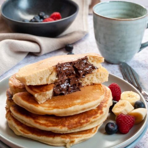 a stack of four large pancakes on a light grey plate with sliced bananas, blueberries and raspberries. The top pancake is cut in half to reveal a molten Nutella filling. A blue cup of coffee and a black bowl of raspberries and blueberries on a beige linen napkin sits in the background.