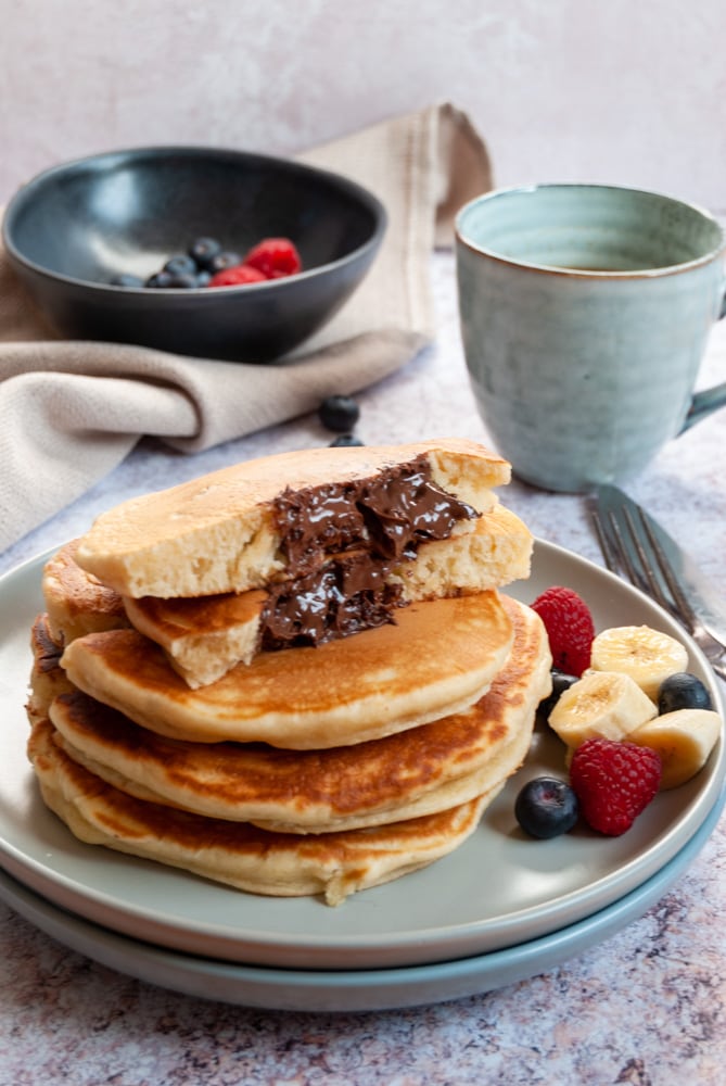 a stack of four large pancakes on a light grey plate with sliced bananas, blueberries and raspberries. The top pancake is cut in half to reveal a molten Nutella filling. A blue cup of coffee and a black bowl of raspberries and blueberries on a beige linen napkin sits in the background.