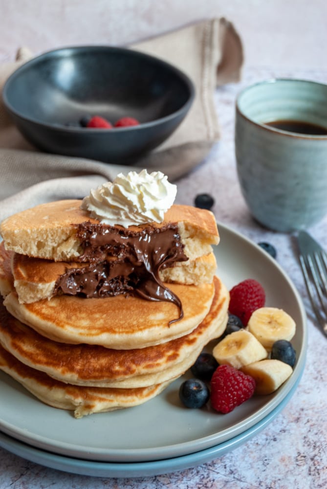 a stack of four pancakes on a light blue plate with sliced bananas, blueberries and raspberries.  The top pancake is broken in half to reveal a molten Nutella middle and is topped with whipped cream.  A blue cup of coffee, a black bowl of raspberries and blueberries and a beige napkin is in the background.