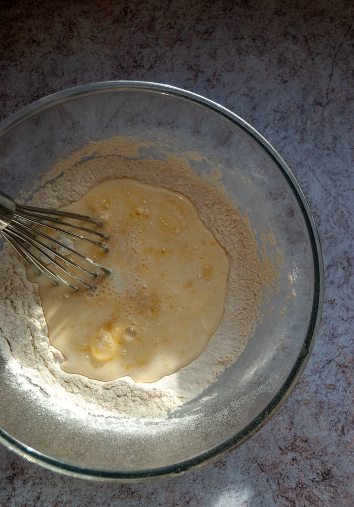 a large glass bowl with flour, eggs and milk being whisked to make a pancake batter on a marbled grey worktop.