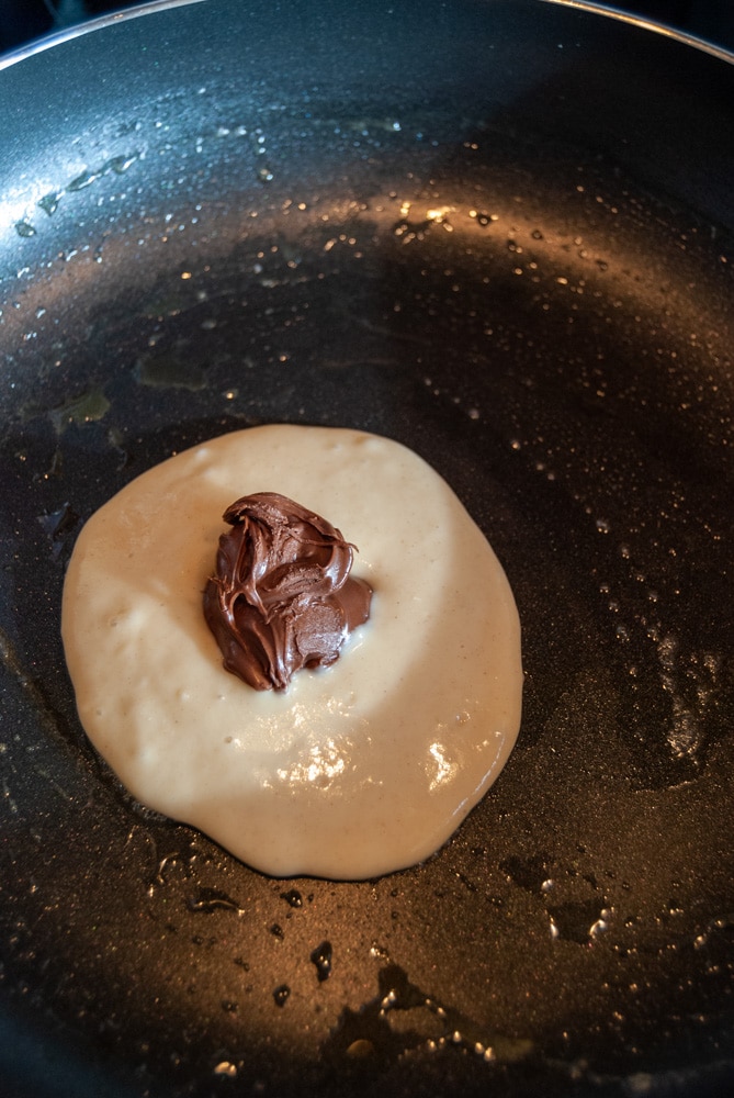 Pancake batter topped with a spoonful of Nutella chocolate spread in a frying pan.